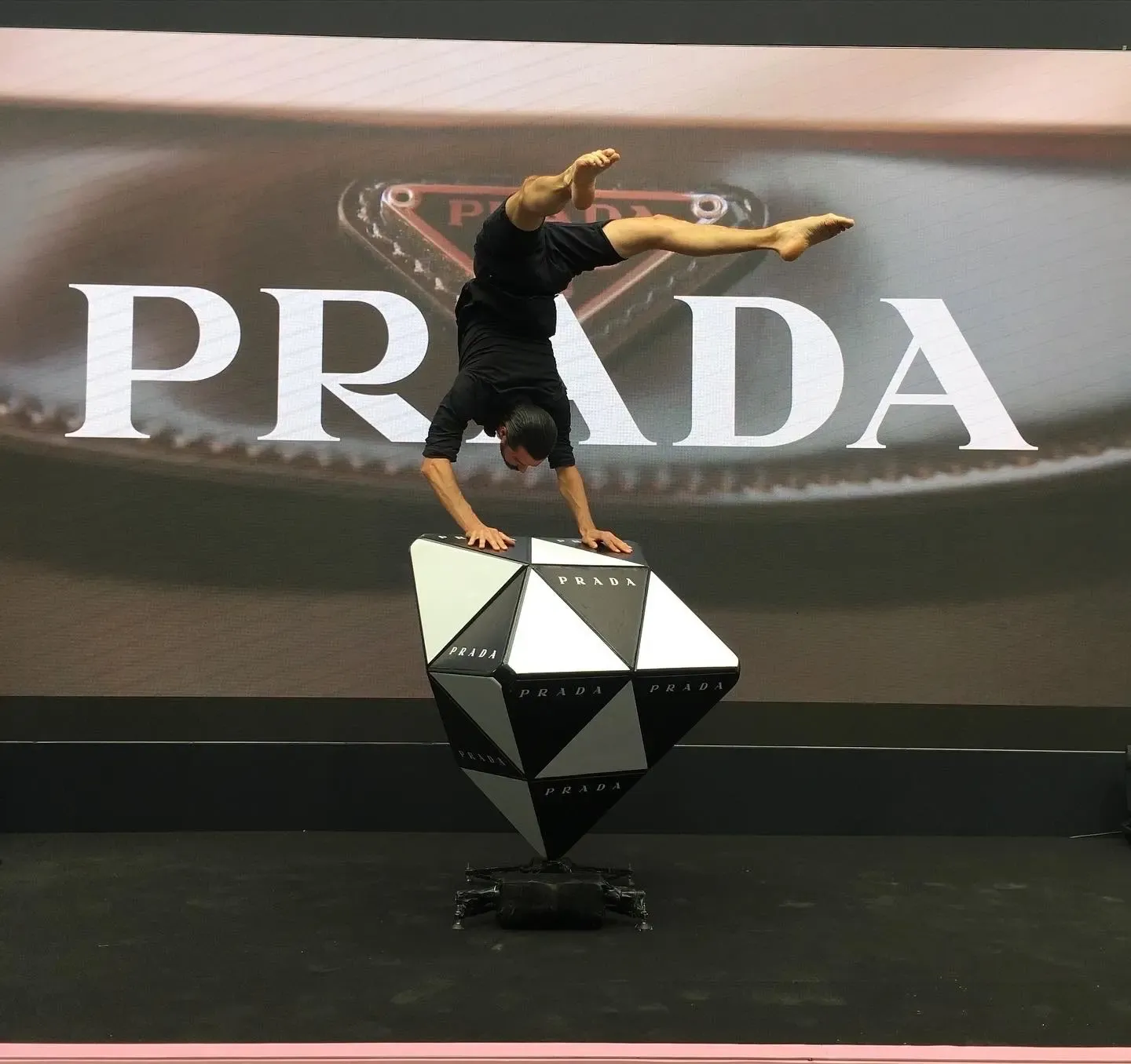 Hand balancer in black doing a split-leg handstand on a giant black-and-white Prada diamond prop, in front of a large Prada logo screen