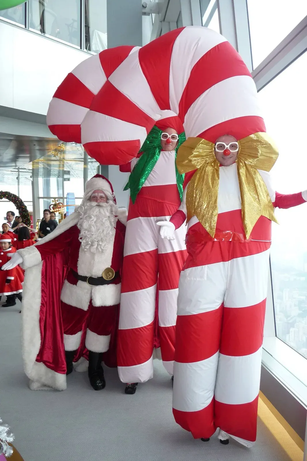 Two inflated candy cane stilt walkers beside Santa, with children in Santa hats behind them
