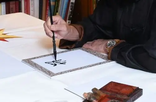 Close-up of a hand writing Chinese characters with a calligraphy brush on paper in a patterned frame, a carved seal box beside it