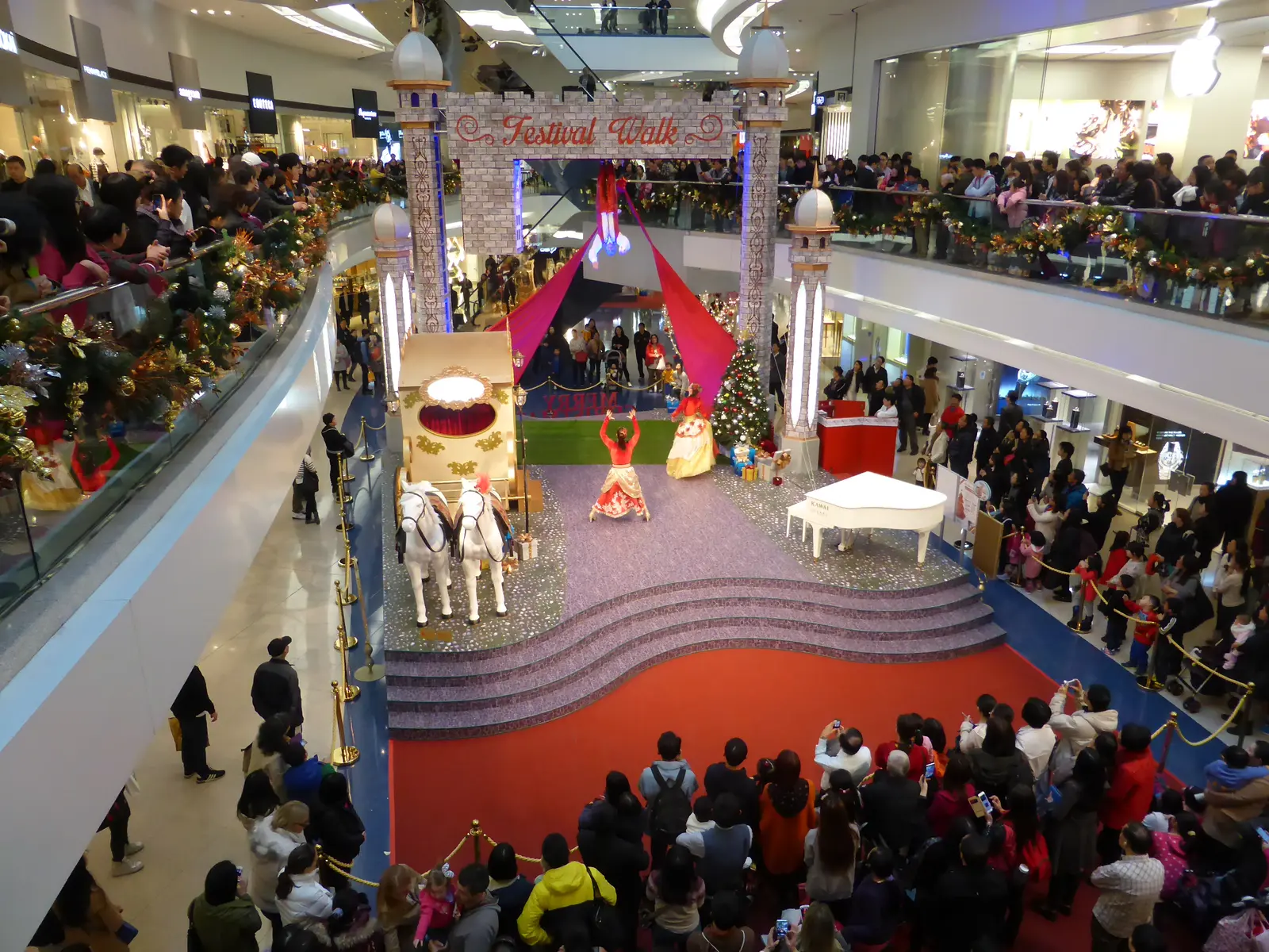 Festival Walk Christmas show from above: an acrobat hangs upside down over dancers, a horse-drawn carriage and the crowd