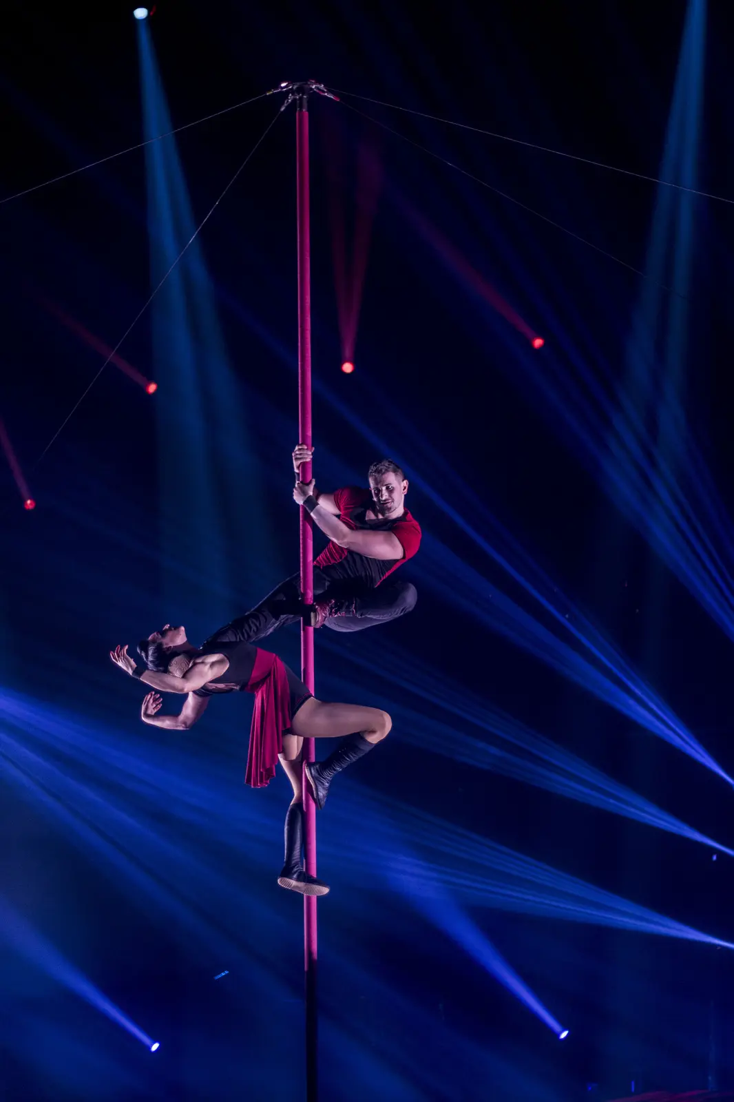 Two acrobats in red and black holding a pose on a vertical Chinese pole under blue stage light