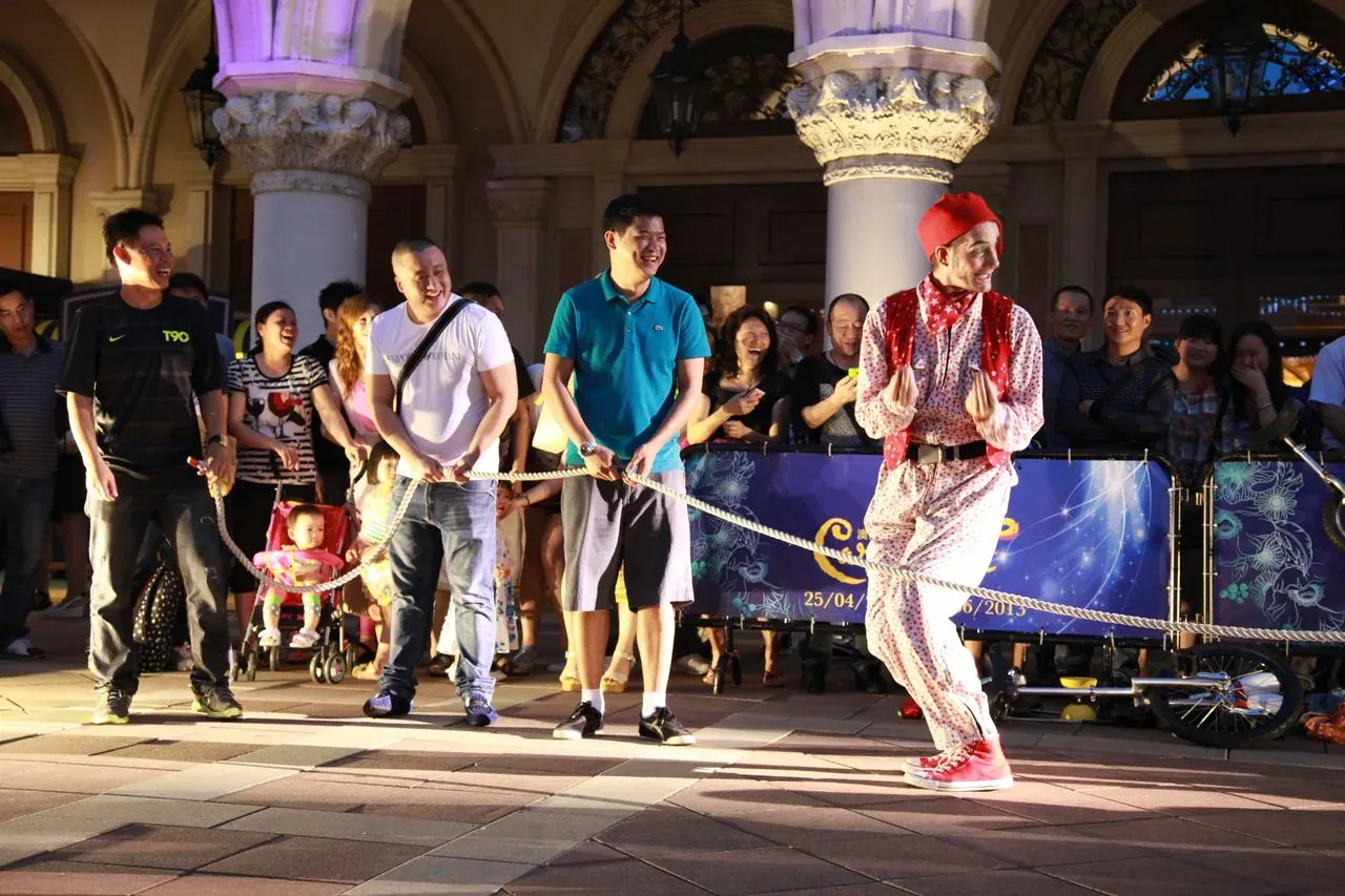 Clown in a red hat and red waistcoat over a polka-dot outfit leading a tug of war, laughing spectators holding the rope, in front of an ornate colonnaded building