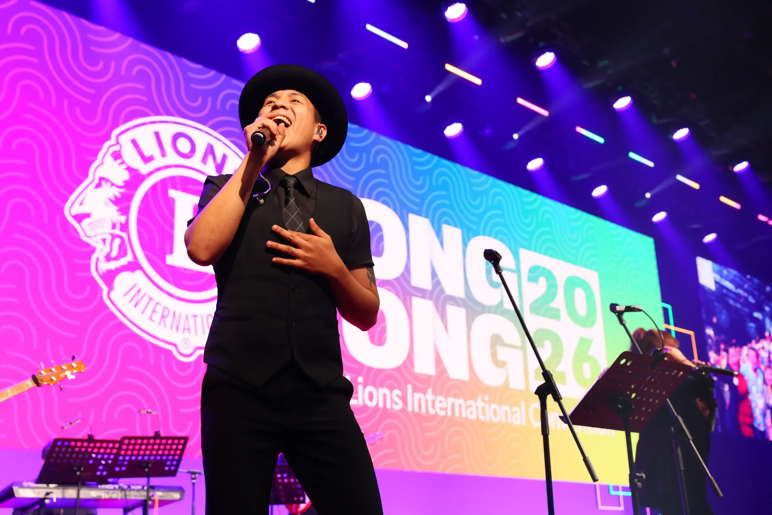Singer in a black hat and waistcoat performing on stage in front of a Lions International Hong Kong 2026 screen