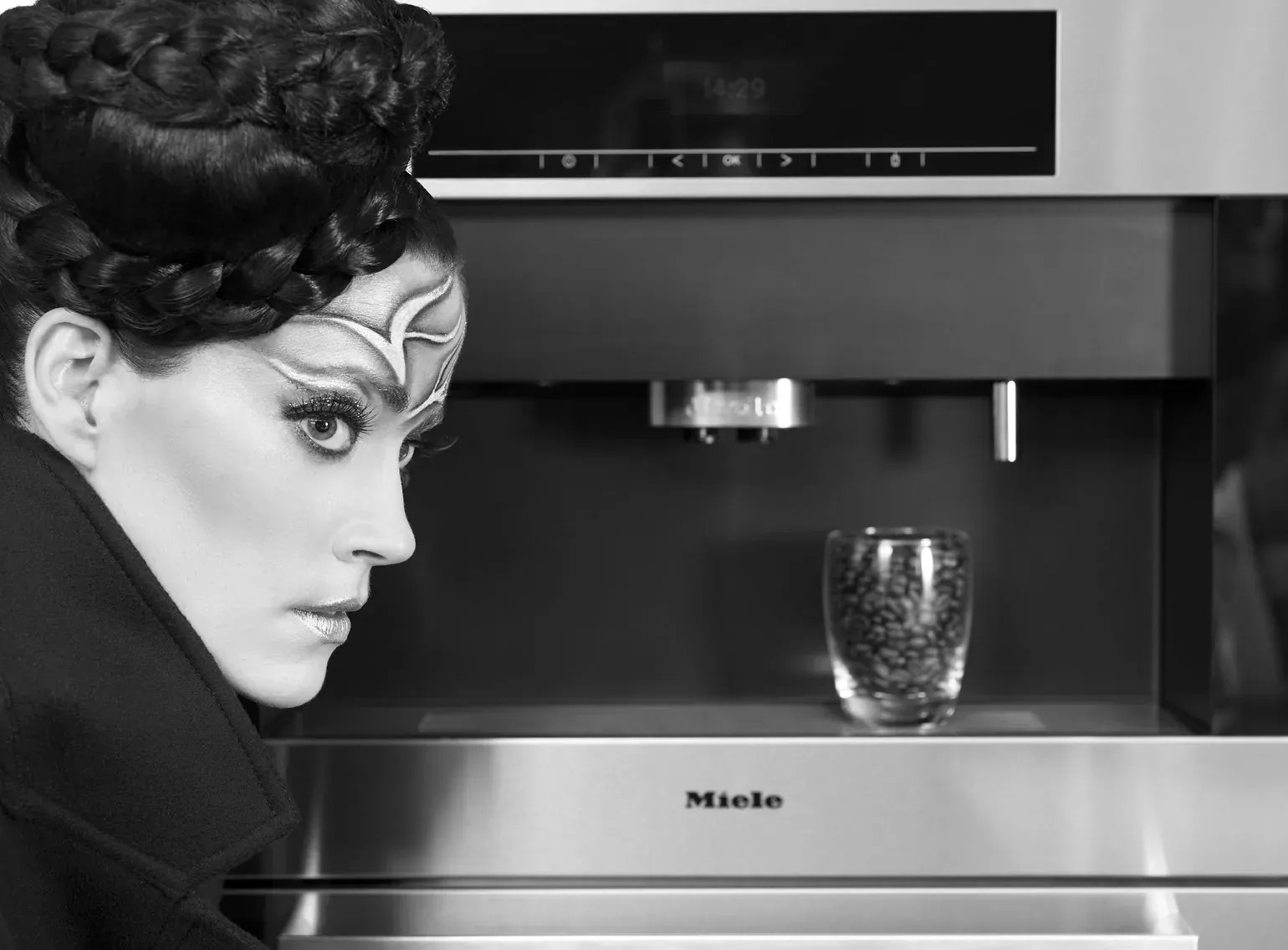Black-and-white photo of a model with a braided crown of dark hair and silver face paint, looking sideways beside a built-in Miele coffee machine with a glass of coffee beans