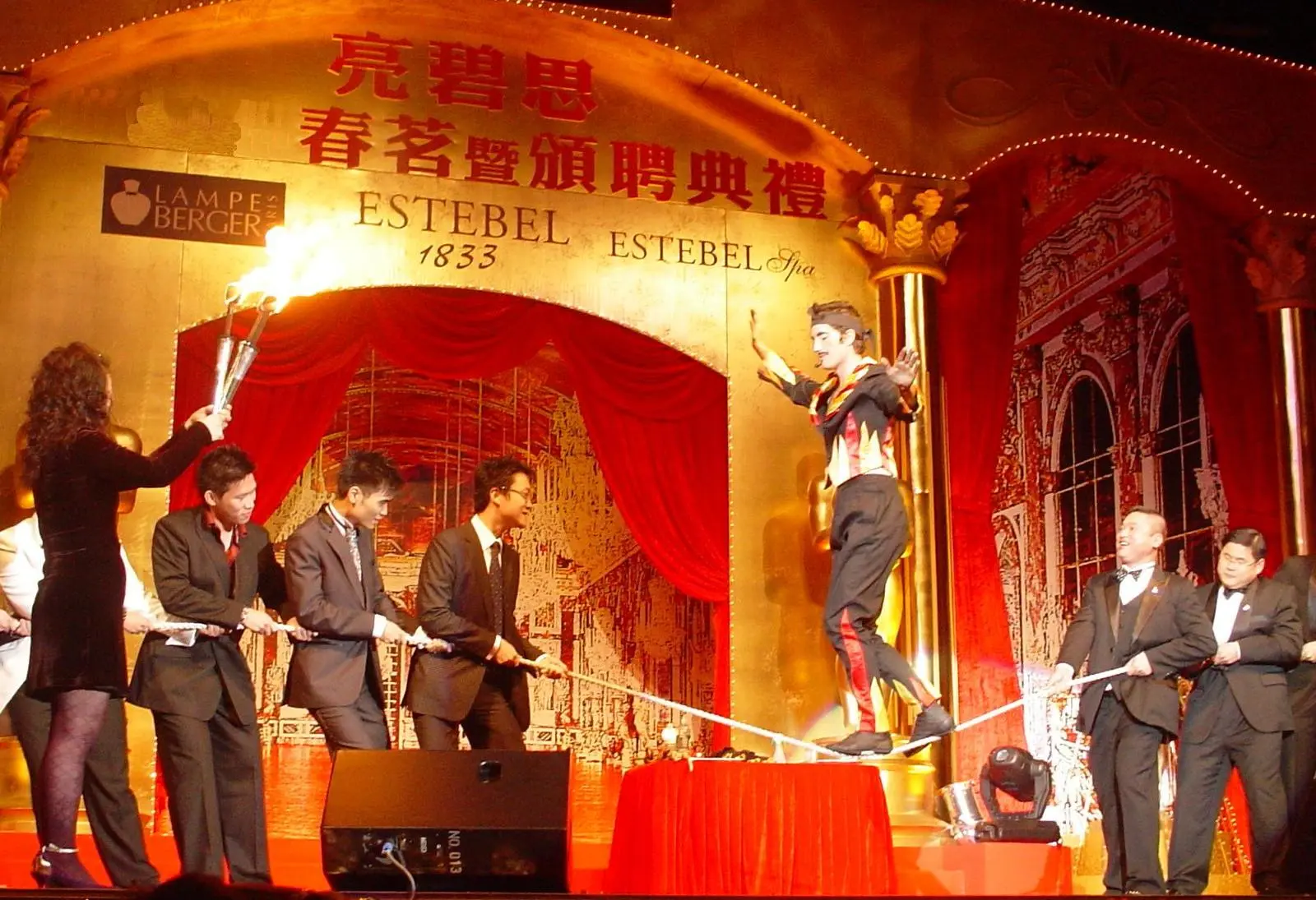 Fire show on a gala stage: a woman holds flaming torches beside a performer walking a rope held by guests