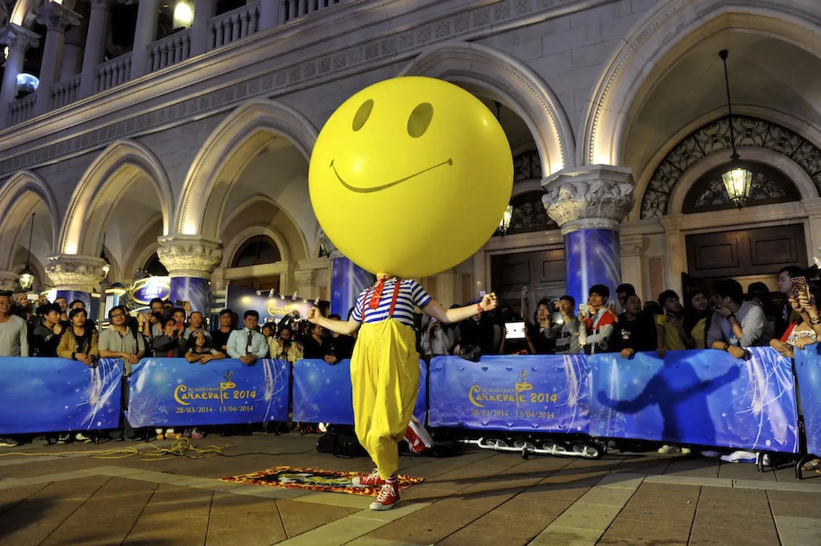 Giant yellow smiley balloon-head street performer in front of a crowd at night