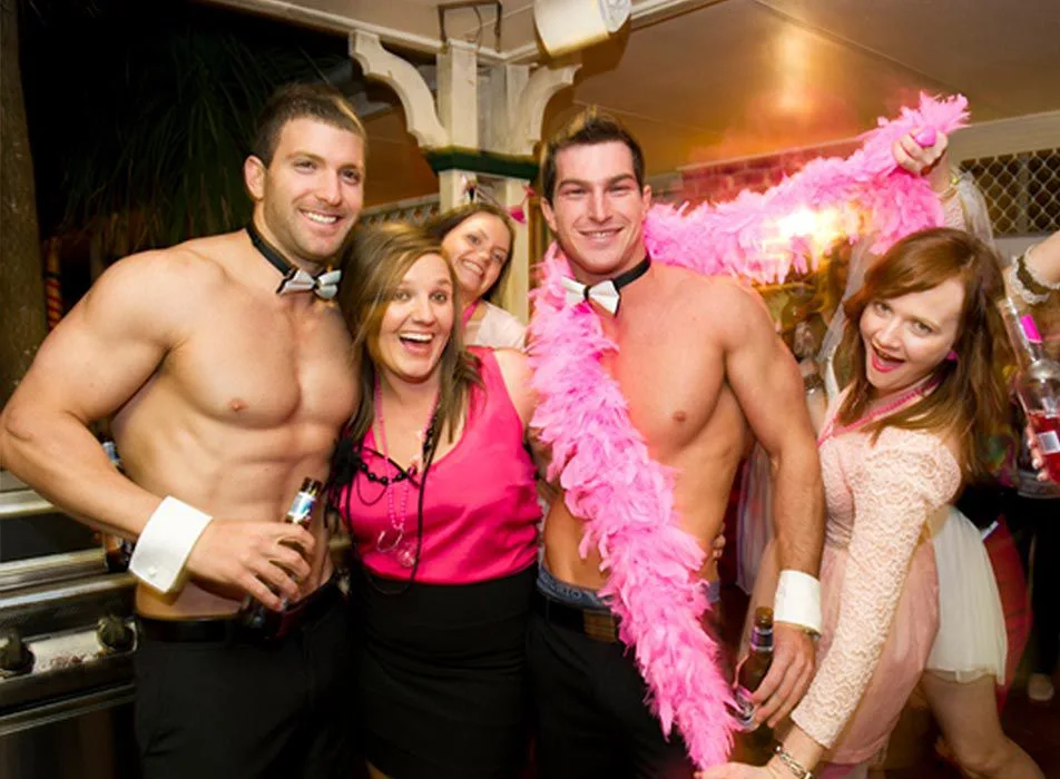 Two shirtless male entertainers in bow-tie collars and cuffs posing with three smiling women at a hen party, a pink feather boa draped over one of the men