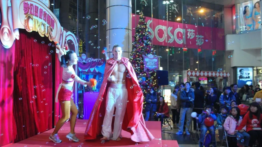 Western performers on a Christmas circus stage in a Hong Kong shopping mall: a performer in a red cape and a dancer in red among bubbles