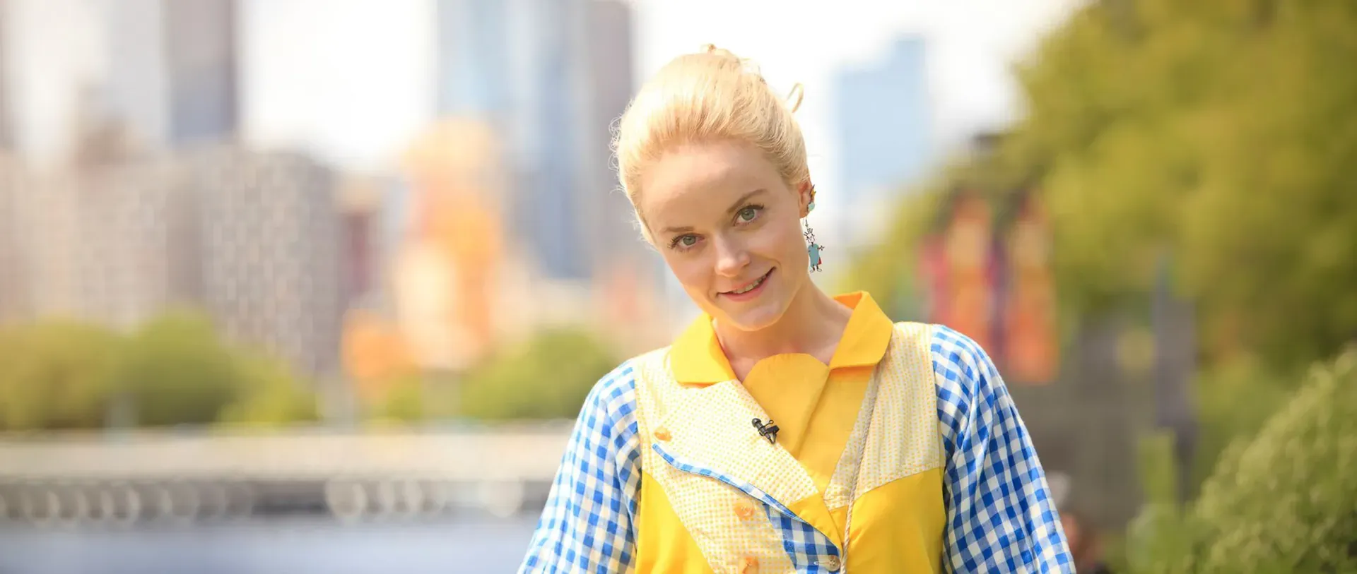 Smiling blonde female MC in a yellow and blue gingham costume with a lapel mic, a city skyline behind her