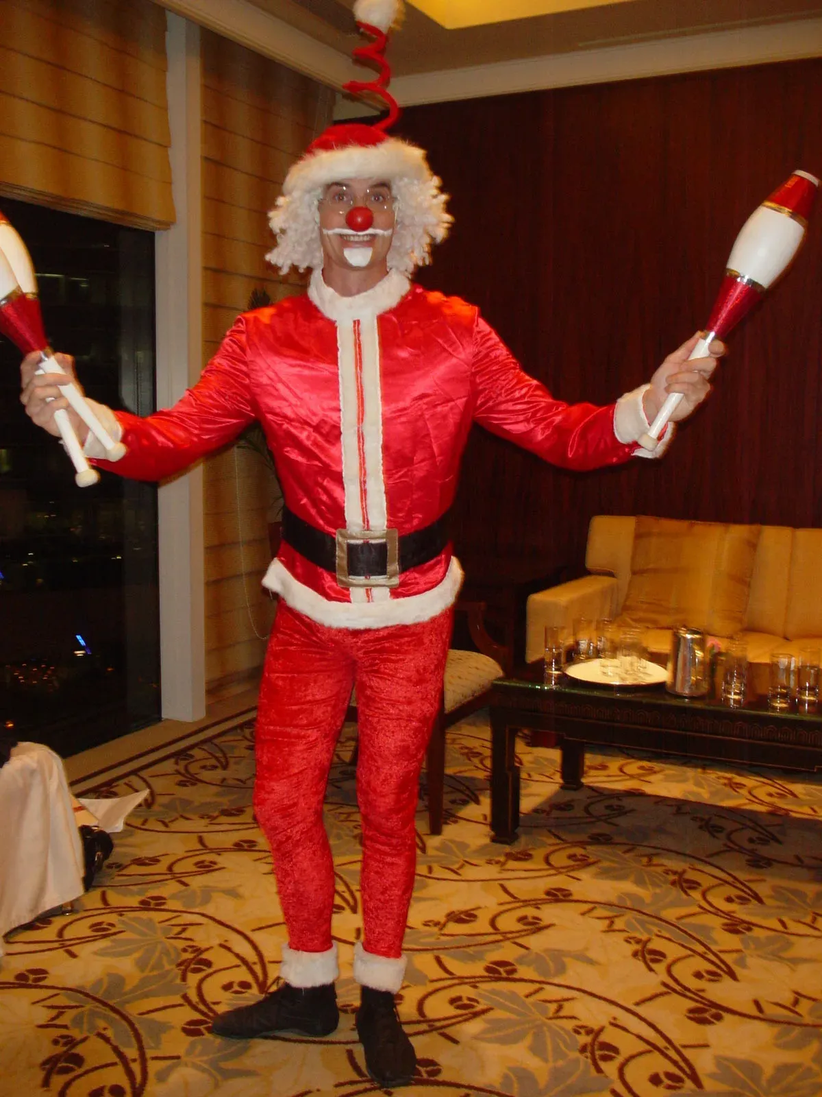 Clown Santa with a red nose and curly white wig juggling clubs in a hotel suite