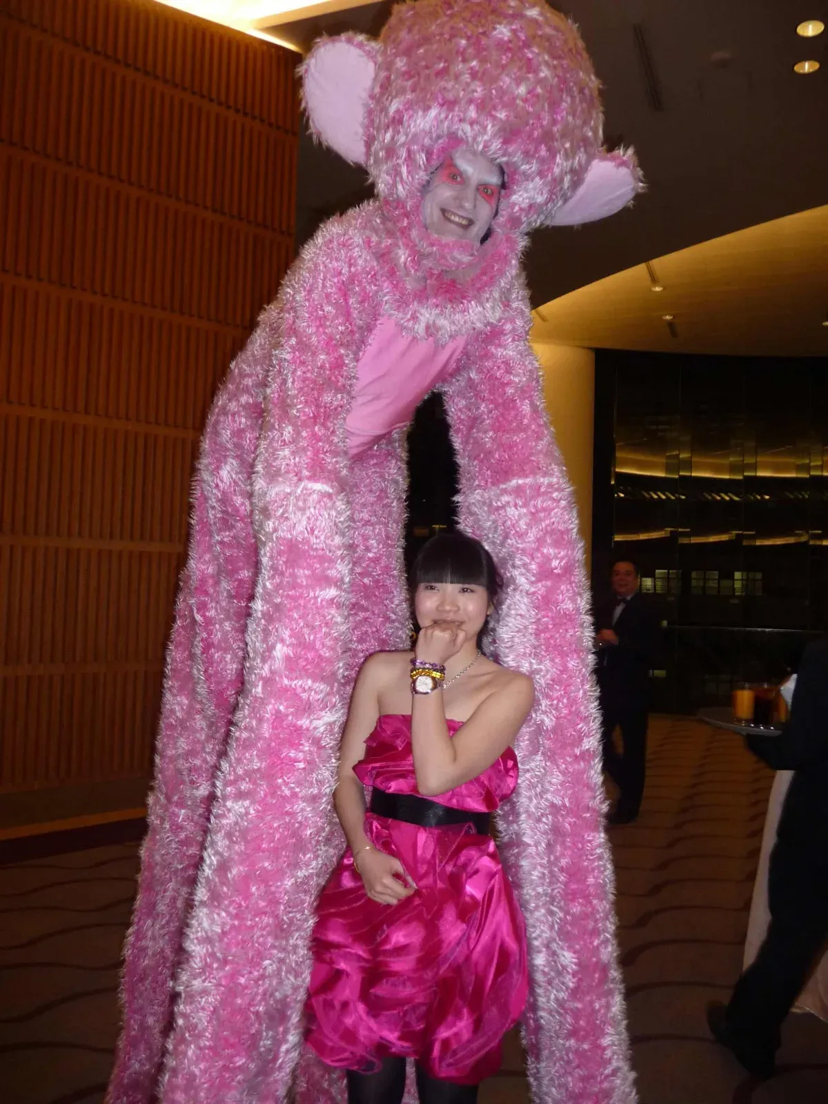 Pink fur animal character on stilts with a woman in a pink dress at a gala dinner