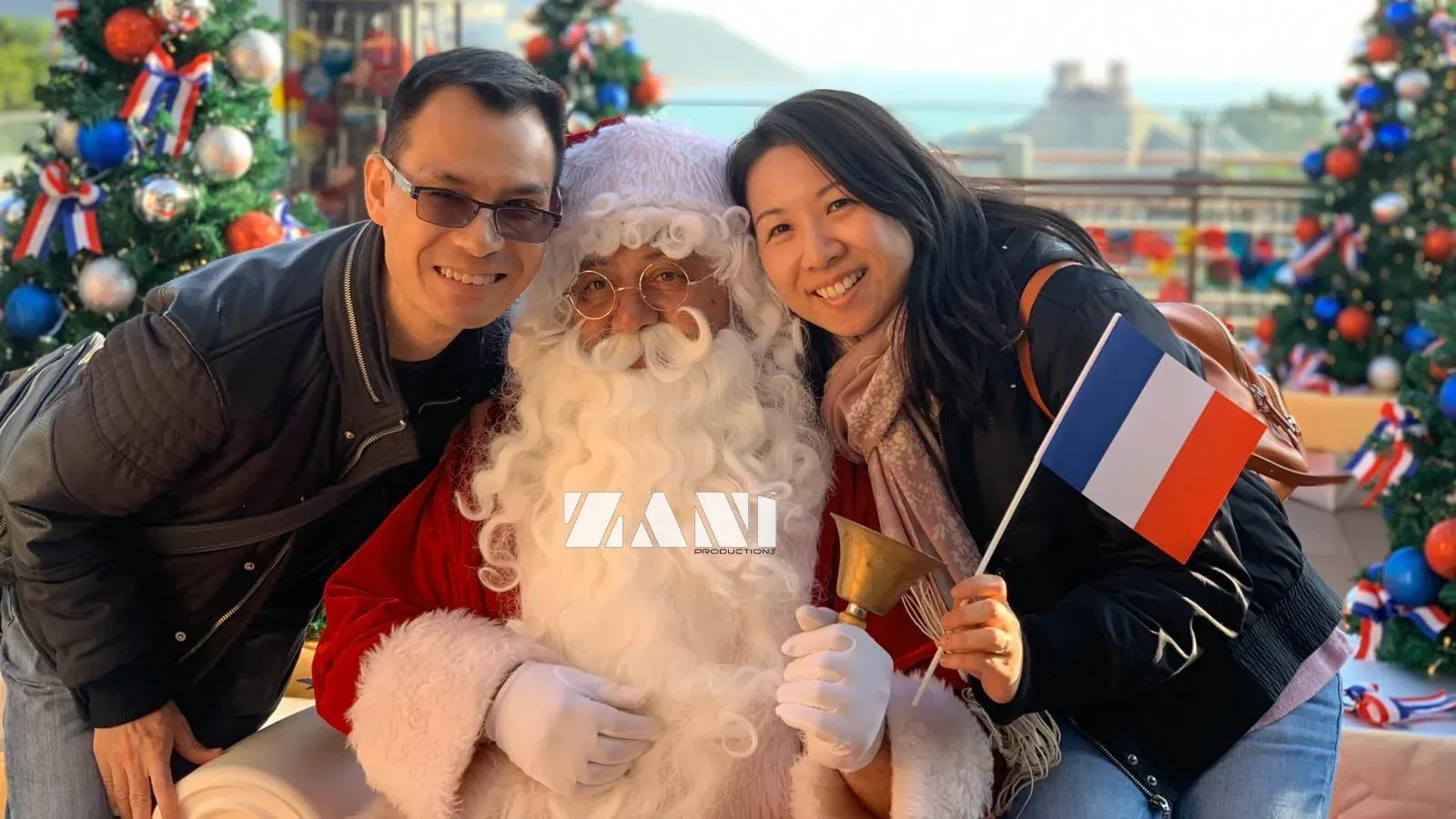 Santa with a hand bell posing with a smiling couple holding a French flag, between Christmas trees