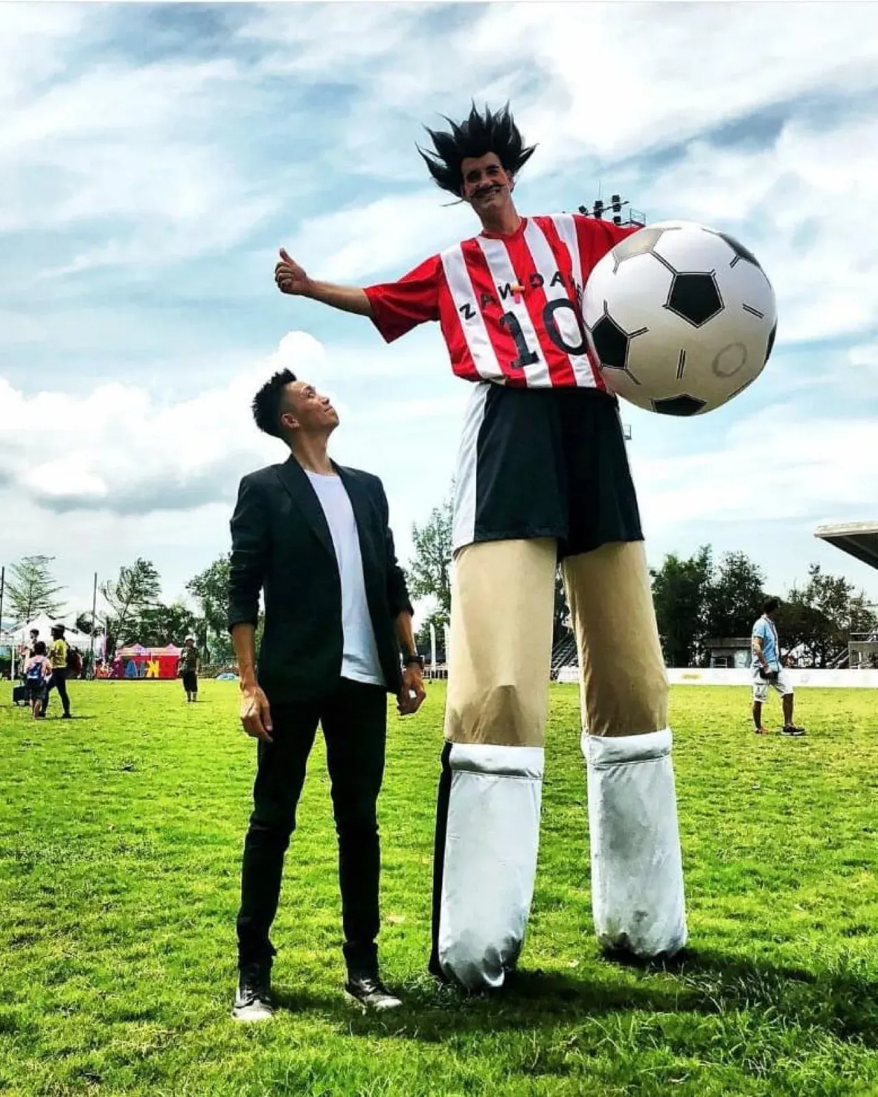 Footballer stilt walker in a red and white striped shirt holding a giant football beside a guest on a lawn