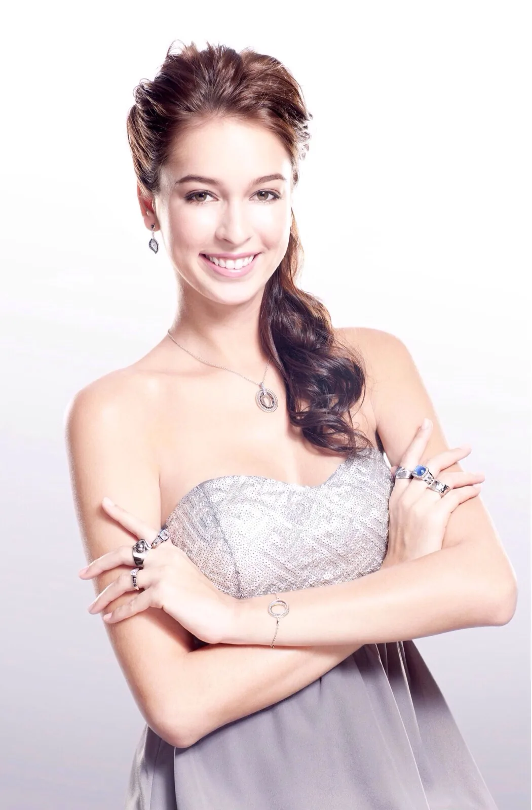 Smiling model with a long side braid in a strapless silver dress, arms crossed to show her rings, bracelet and pendant, white studio background
