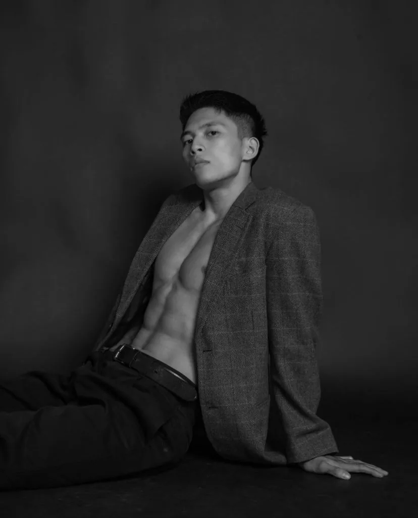 Black and white studio photo of a young Asian male model sitting on the floor in an open checked tweed blazer with no shirt, dark trousers and a belt