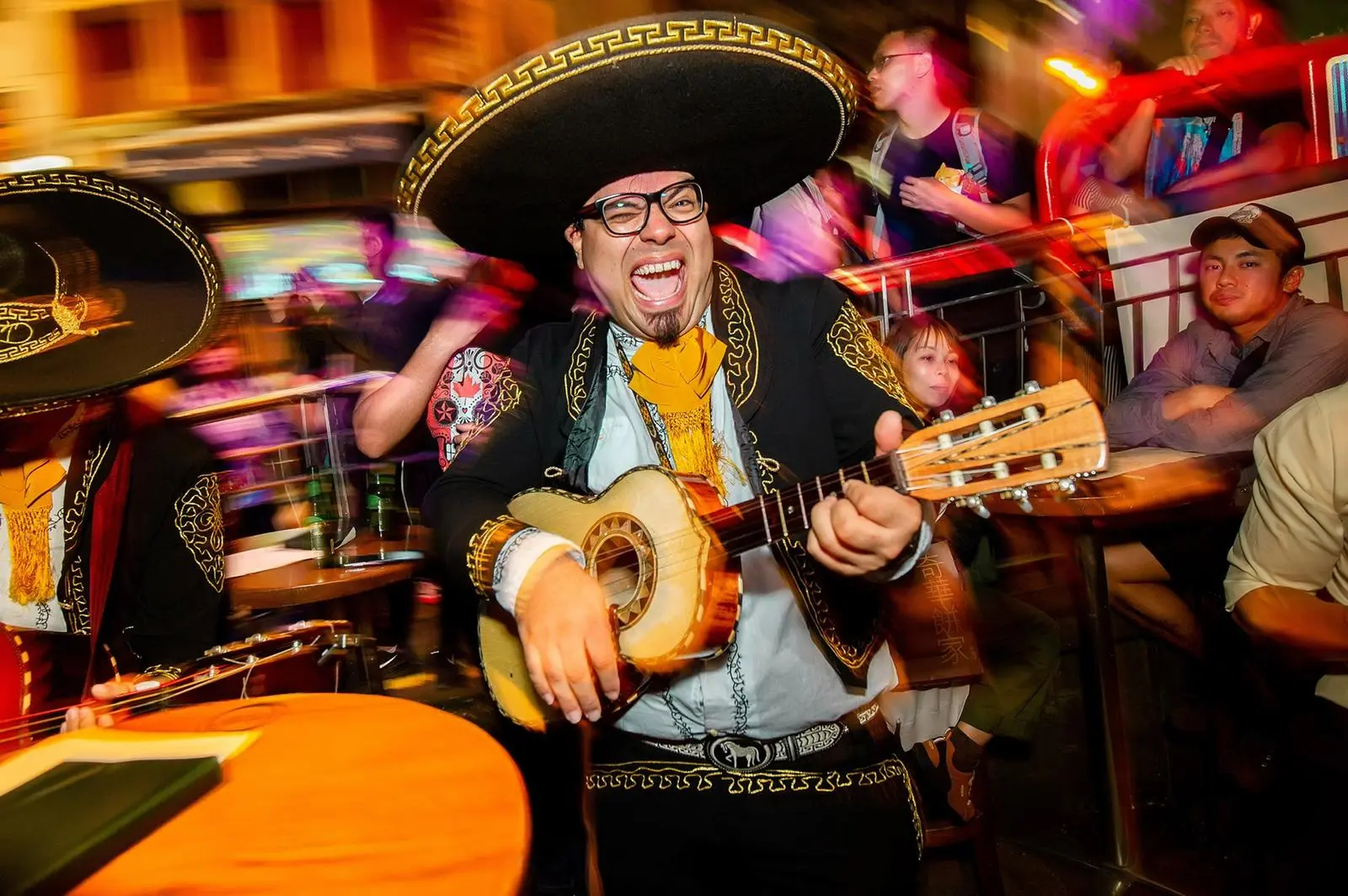 Mariachi guitarist in a black sombrero, glasses and a yellow scarf singing and playing guitar at a night-time party