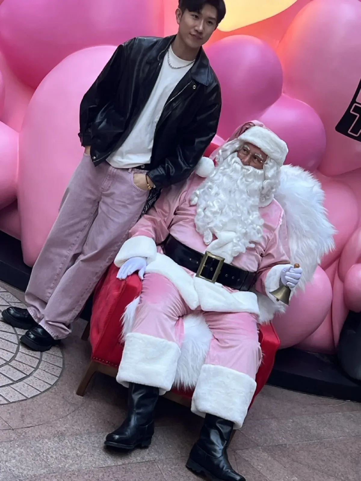 Pink Santa with a hand bell seated on a red chair beside a young man in a black leather jacket, pink balloons behind