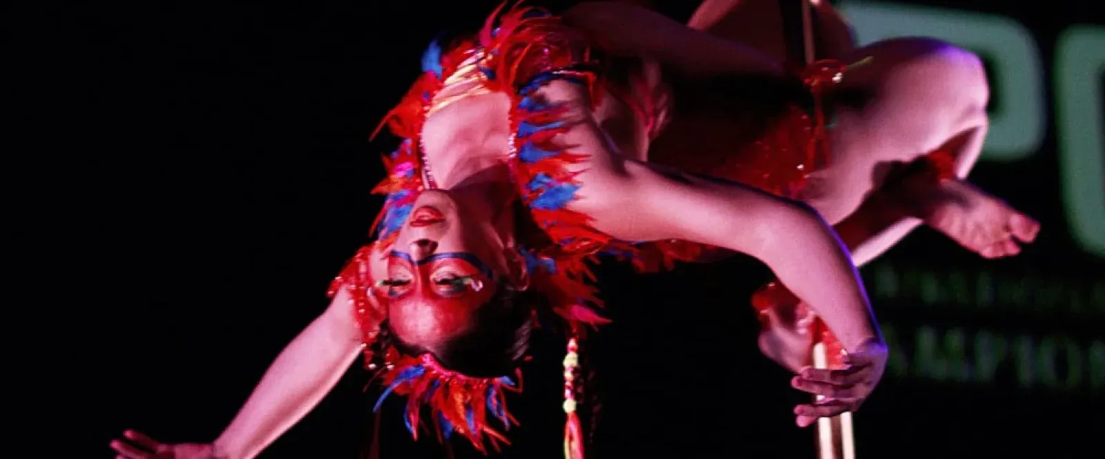 Pole dancer in a red feathered costume hanging upside down from a pole under red light