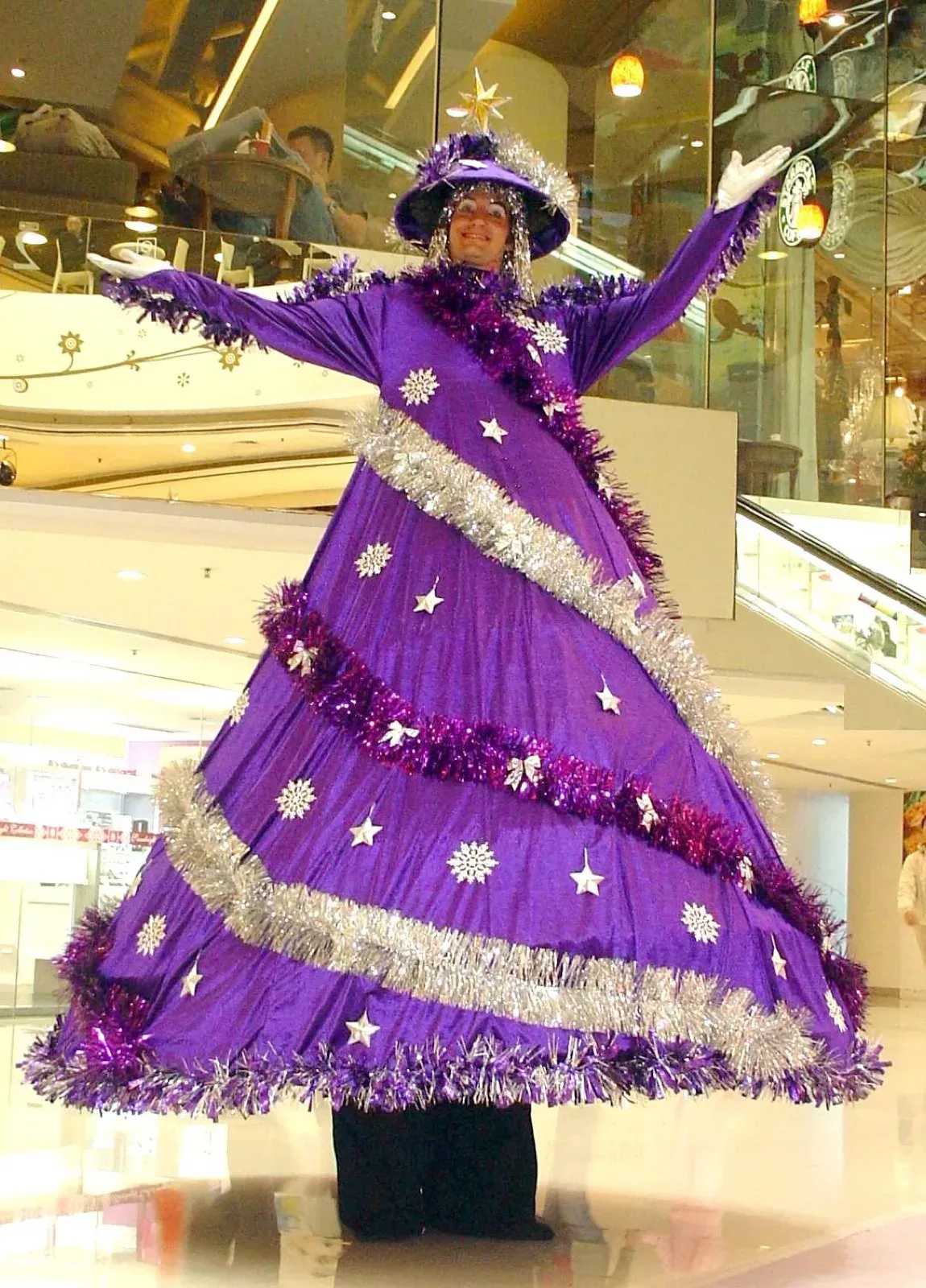Performer on stilts in a purple Christmas tree costume with silver tinsel, arms raised, in a shopping mall