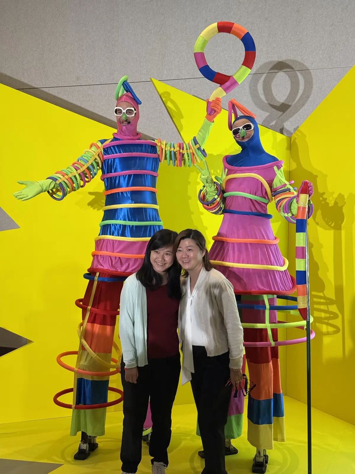Two stilt walkers in striped rainbow costumes covered in coloured hoops, posing with two women in front of a yellow backdrop