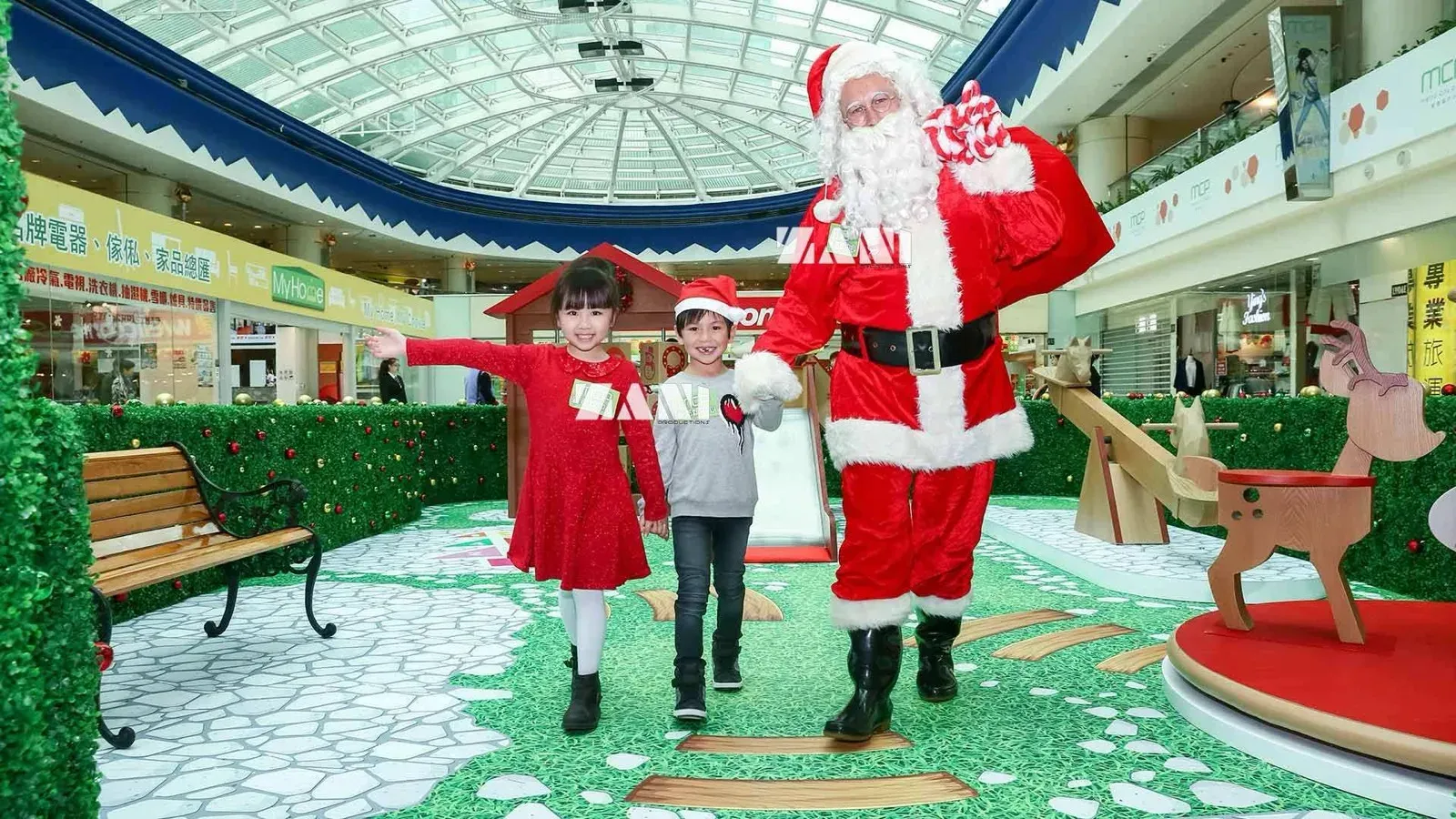 Santa with his sack walking with two children through a garden-themed Christmas set with hedges and wooden reindeer