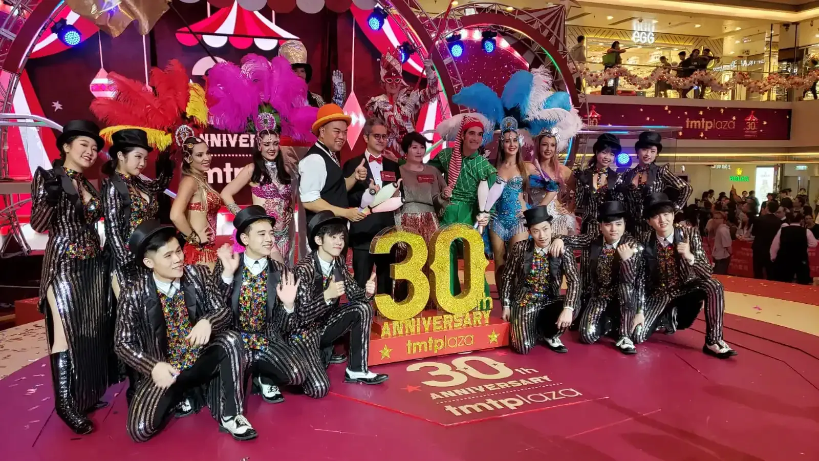 Show cast in striped sequin suits, feathered headdresses and costumes posing on stage at the tmtplaza 30th anniversary in a shopping mall