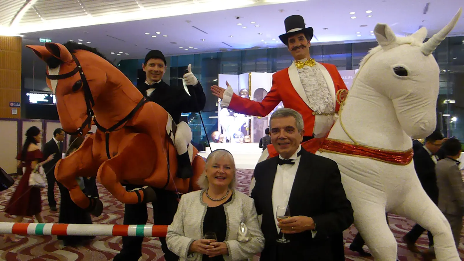 Two performers riding costume horses, a brown show-jumping horse and a white unicorn, posing with two guests in evening dress at a gala