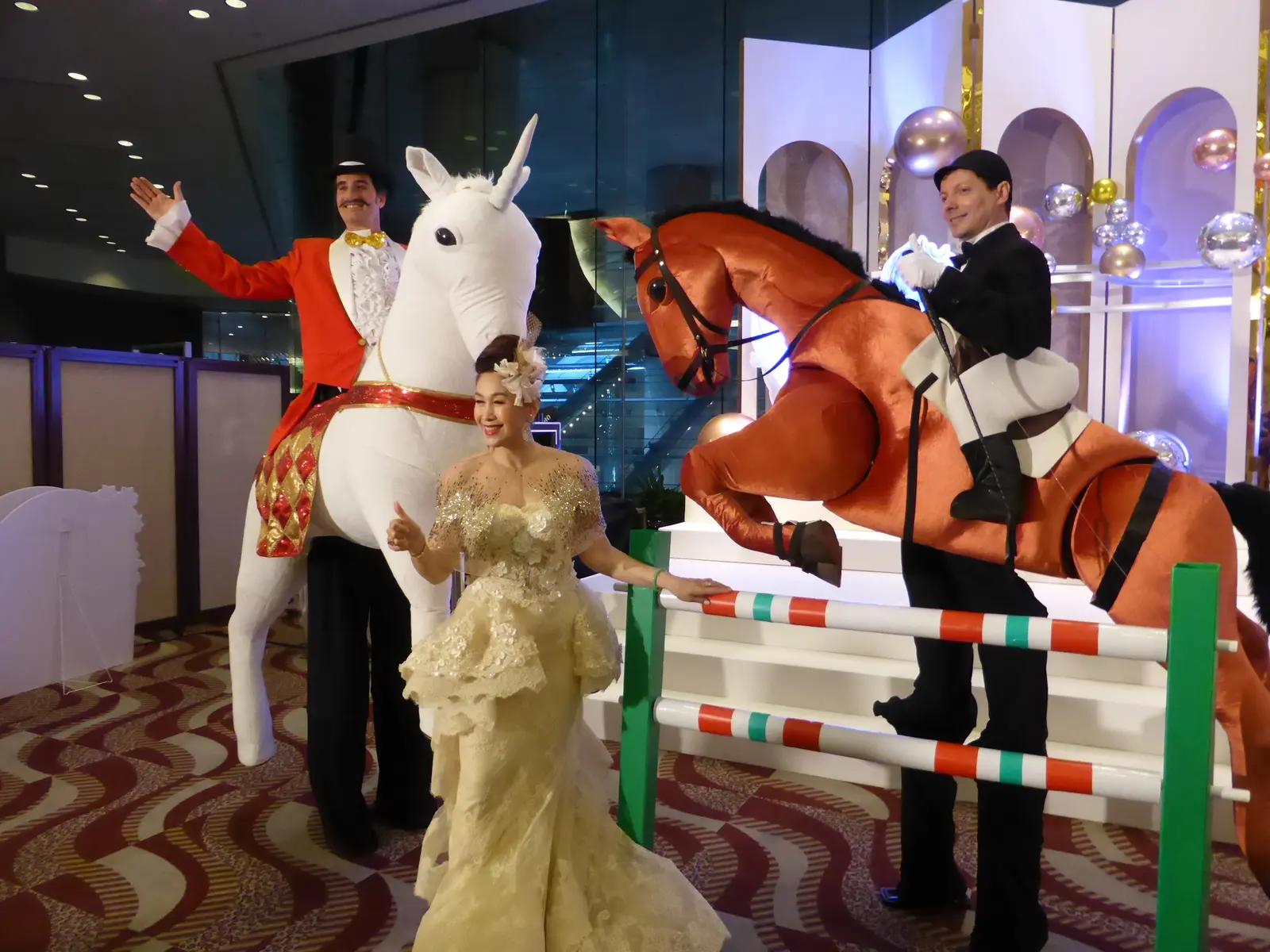 Performers riding a white unicorn and a brown show-jumping costume horse, posing with a guest in a gold lace gown