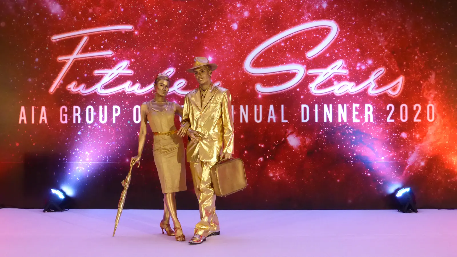 A woman in a gold dress holding a closed umbrella and a man in a gold suit and hat holding a suitcase, standing in front of a red Future Stars backdrop