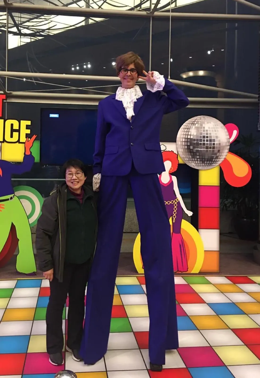 Austin Powers lookalike on stilts in a purple suit beside a guest on a disco-themed stage