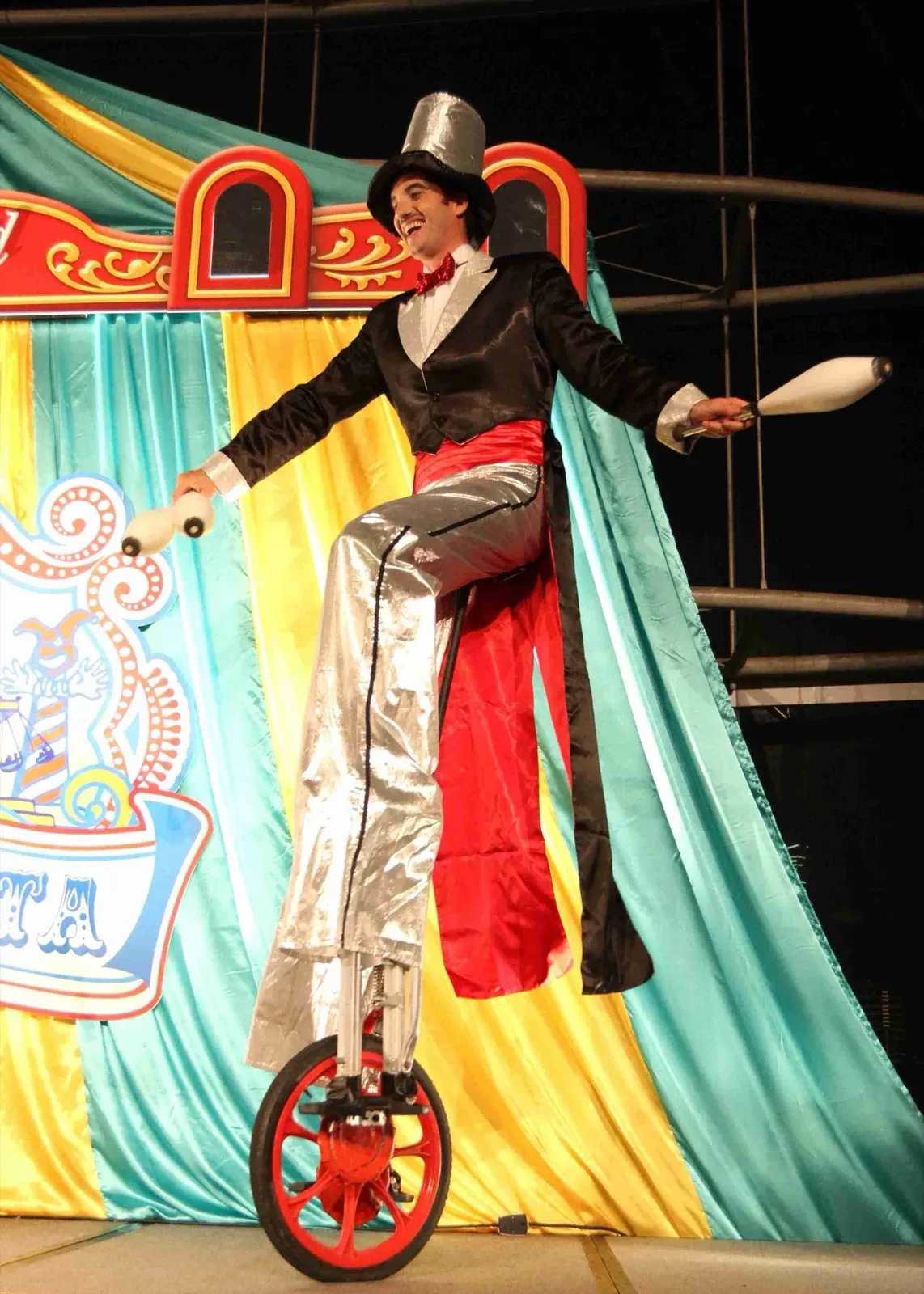 Performer in a top hat and tailcoat juggling clubs on a tall unicycle on a circus stage