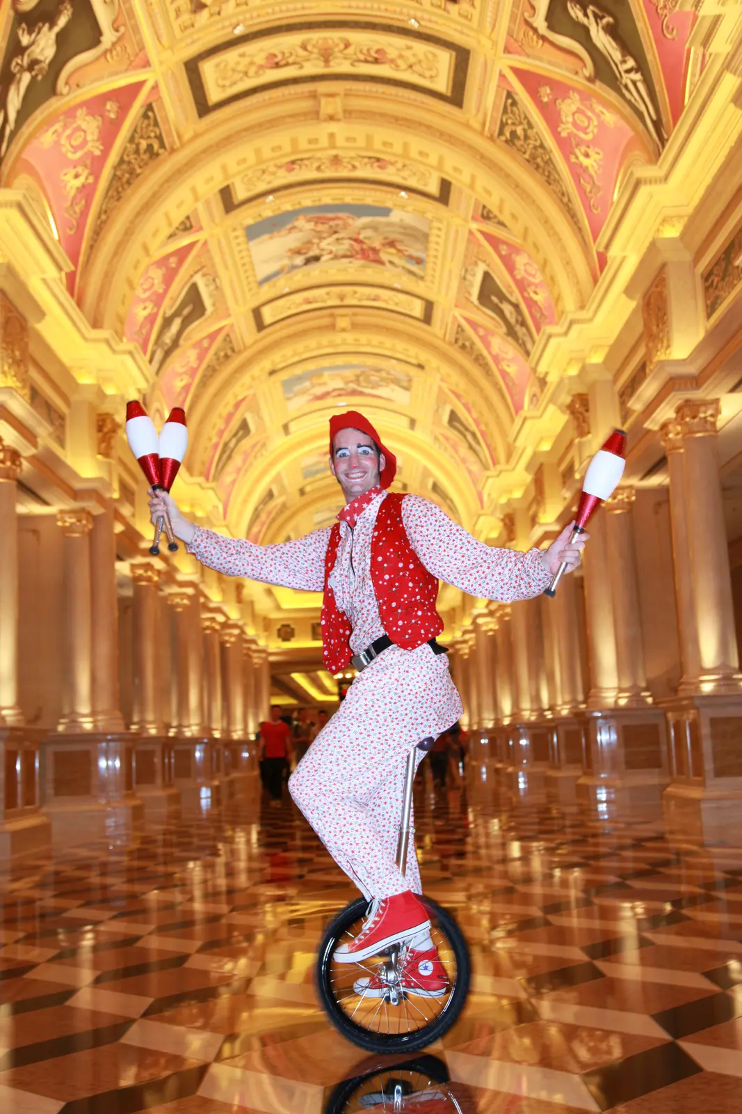 Performer in a red waistcoat and white trousers juggling on a unicycle in an ornate gold Venetian-style hall